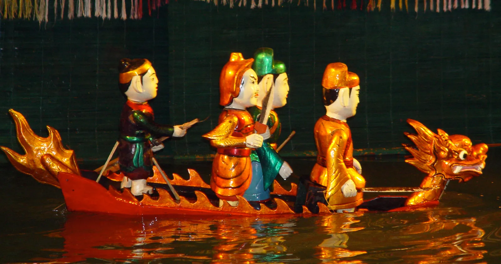 Four painted wooden puppets in gold, orange, teal and green costumes riding a red dragon-headed boat on dark water; a traditional water-puppet show in Hanoi, not the Nha Trang show