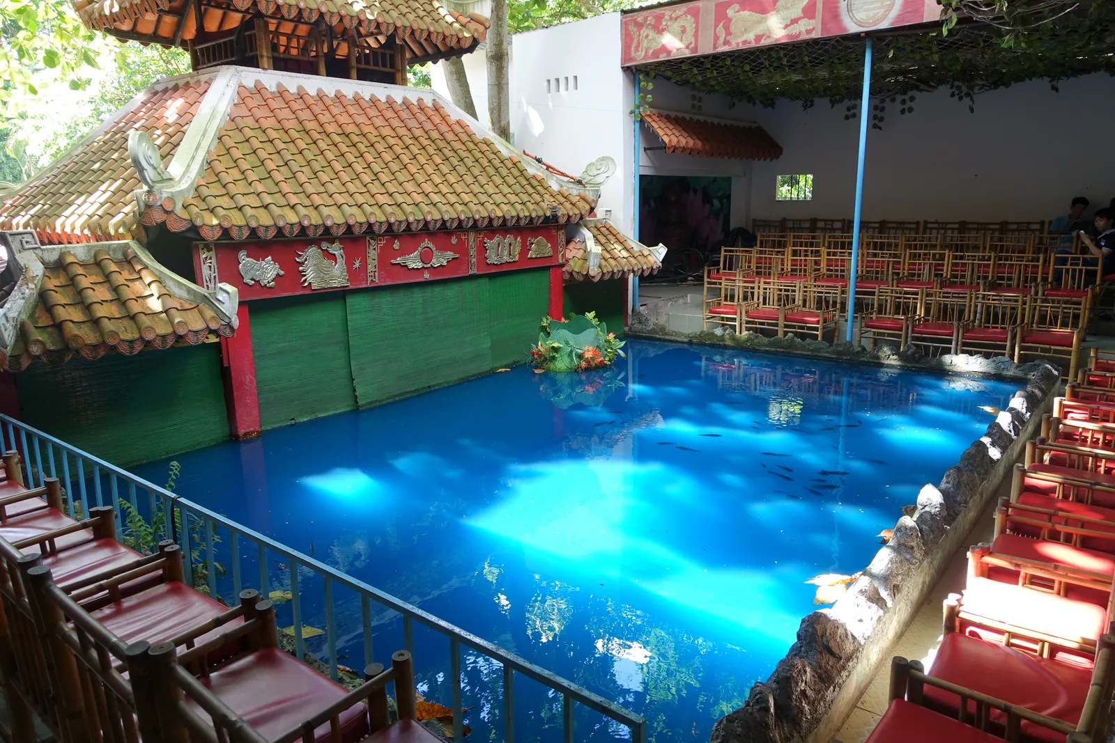 A pool lit bright blue in front of a red-and-green pagoda-style pavilion with tiled roofs, with rows of red seats along the side, at a museum in Ho Chi Minh City