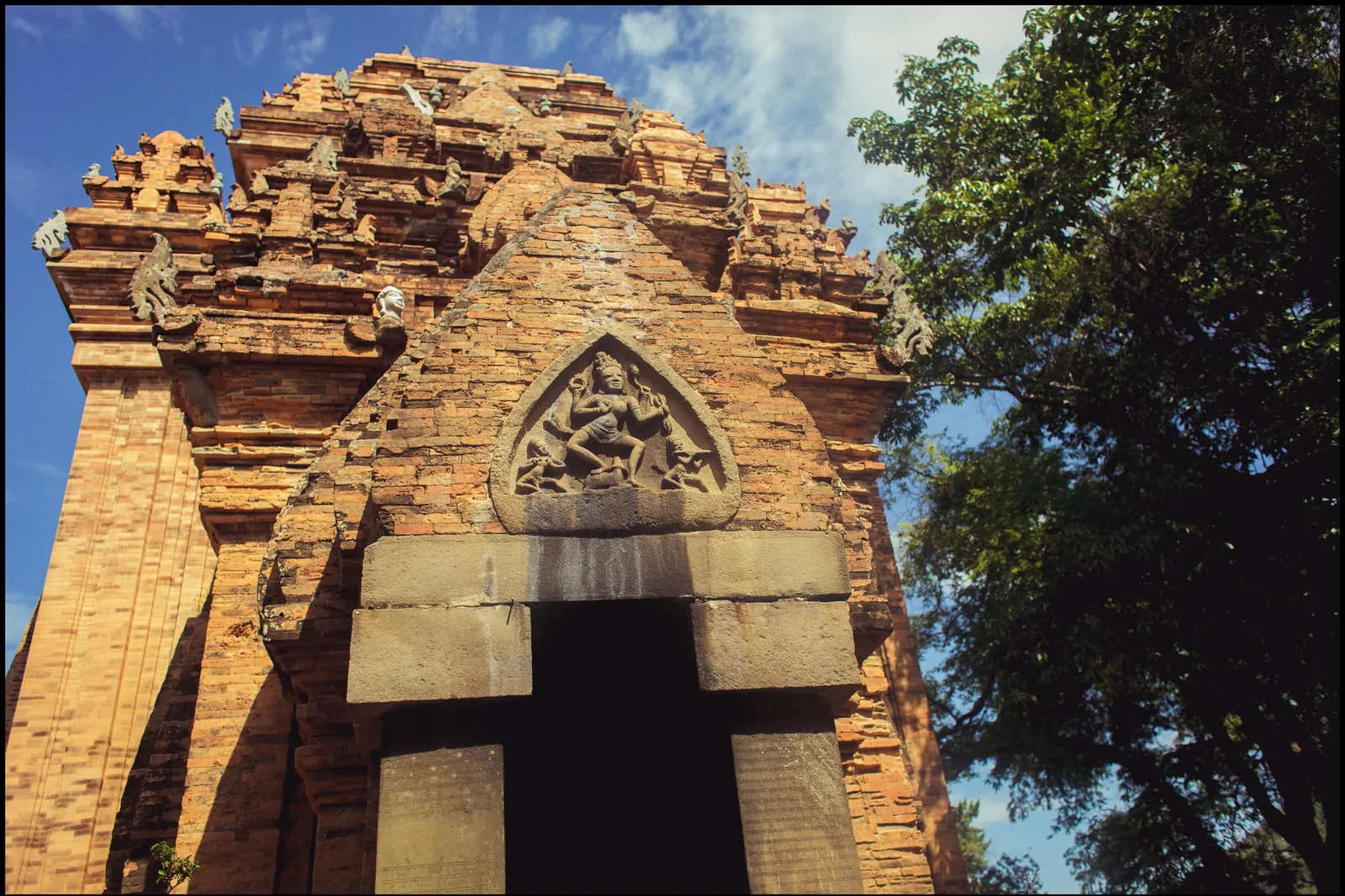 A weathered brick Cham tower with a carved arched pediment above a stone doorway, seen from below against a blue sky; Po Nagar in Nha Trang
