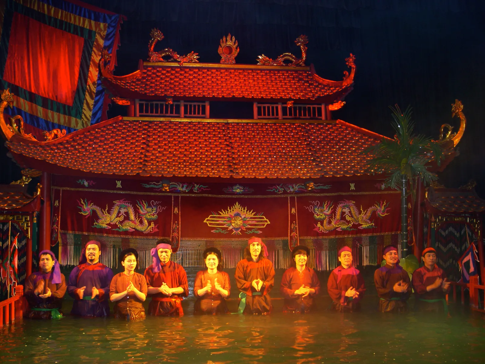 A line of puppeteers in rust-red clothes standing waist-deep in water in front of a red-roofed pagoda-style stage, hands pressed together in thanks; a traditional water-puppet show in Hanoi